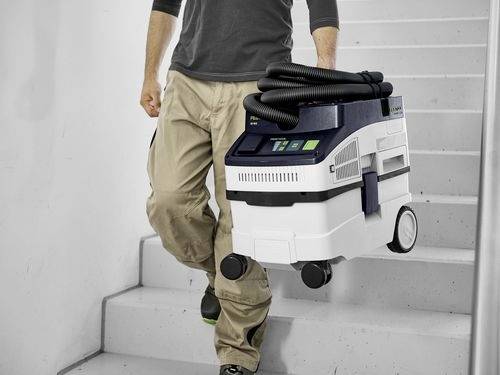 Eine Person trägt einen kompakten Staubsauger eine Treppe hinunter, wobei dessen Schlauch ordentlich auf der Oberseite aufgerollt ist. Der Staubsauger verfügt über einen stabilen Griff und Räder.