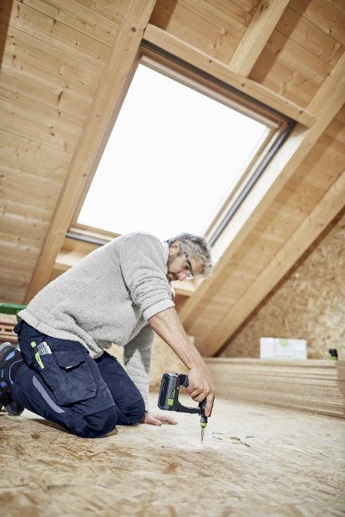 Eine Person kniet auf einem Holzboden unter einem Oberlicht und verwendet eine Bohrmaschine. Die Umgebung scheint ein Dachboden oder Loftraum im Bau zu sein.