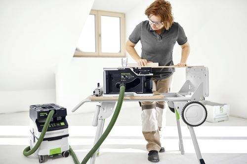 Eine Person bedient eine kompakte Tischkreissäge, die an einen Staubabsauger in einer hellen Werkstatt angeschlossen ist und sich darauf konzentriert, ein Holzbrett zu schneiden.