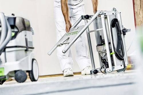 Eine Person in weißer Arbeitskleidung richtet eine Säge in einer Werkstatt ein, während im Hintergrund Werkzeuge und Ausrüstung zu sehen sind.
