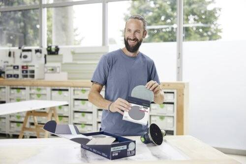 Eine Person in einer Werkstatt lächelt, während sie eine Packung Schleifscheiben neben einer offenen Kiste präsentiert, mit Werkzeugen, die im Hintergrund sichtbar sind.