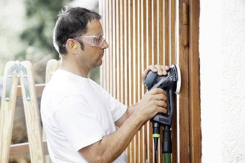 Eine Person verwendet eine elektrische Schleifmaschine, um eine Holzoberfläche zu glätten, trägt dabei Schutzbrille und Gehörschutz, während im Hintergrund eine Leiter steht.