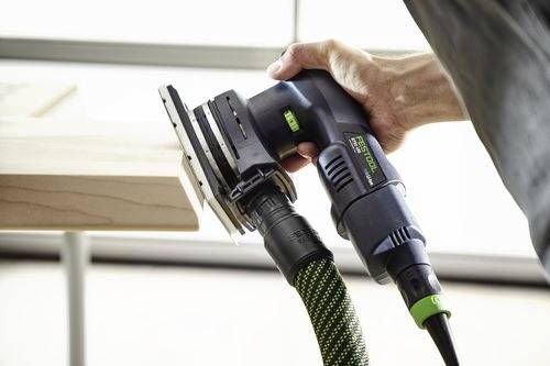 Eine Person benutzt einen elektrischen Schleifer auf einer Holzoberfläche und hält das Werkzeug mit einer Hand. Der Schleifer ist mit einem grün-schwarzen Schlauch verbunden.