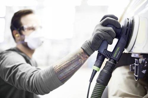 Eine Person mit Sicherheitsbrille und Maske verwendet einen Exzenterschleifer auf der Oberfläche eines Fahrzeugs und betont dabei Sicherheit und Präzision bei der Autoarbeit.