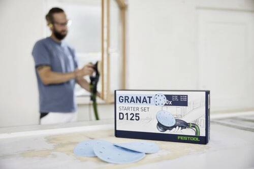 Karton mit der Aufschrift 'Granat D125 Starter Set' mit Schleifscheiben auf einem Tisch. Eine Person im Hintergrund hält ein Werkzeug, leicht unscharf.