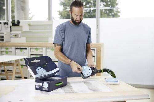 Mann in einer Werkstatt, der ein elektrisches Schleifwerkzeug auf einer Werkbank benutzt, mit einer Box mit Schleifscheiben in der Nähe.