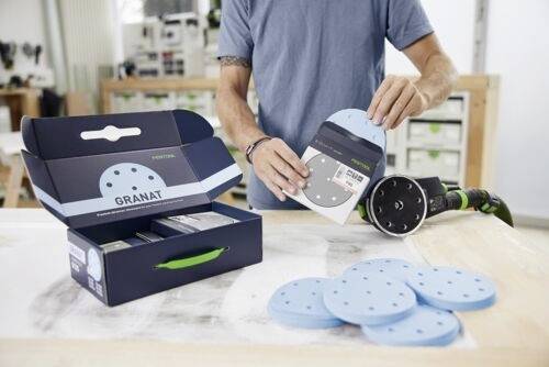 Eine Person in einer Werkstatt hält eine kreisförmige Schleifscheibe neben einer Maschine. Eine mit ‚Granat' beschriftete Box und weitere Scheiben liegen auf einem Tisch.