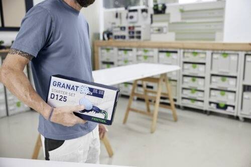 Eine Person in einer Werkstatt hält eine 'GRANAT Starter Set D125' Box. Im Hintergrund sind Regale mit sortierten Behältern zu sehen.