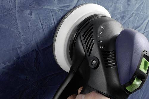 Eine Person verwendet einen elektrischen Schleifer, um eine strukturierte blaue Wand zu glätten. Das Werkzeug ist mit ‚ROTEX' gekennzeichnet.