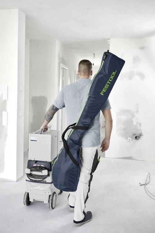 Eine Person, die eine große Festool-Tasche trägt und Ausrüstung auf einem Wagen schiebt, geht durch einen teilweise renovierten, weißwandigen Raum.