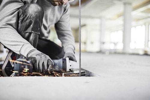 Eine Person in Arbeitskleidung benutzt eine Trennschleifmaschine, um ein Betonstabeisen auf einer Baustelle zu schneiden, wobei Funken in der Umgebung herumfliegen.