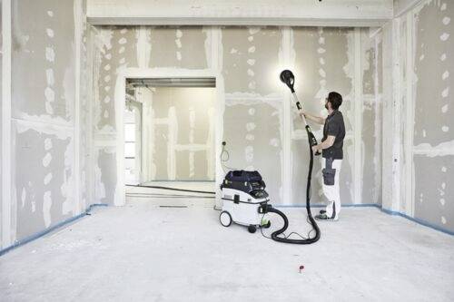 Eine Person in Arbeitskleidung nutzt ein Schleifwerkzeug, das an einen Staubsauger angeschlossen ist, an einer Trockenbauinnenwand und bereitet den Raum für das Malen oder Fertigstellen vor.