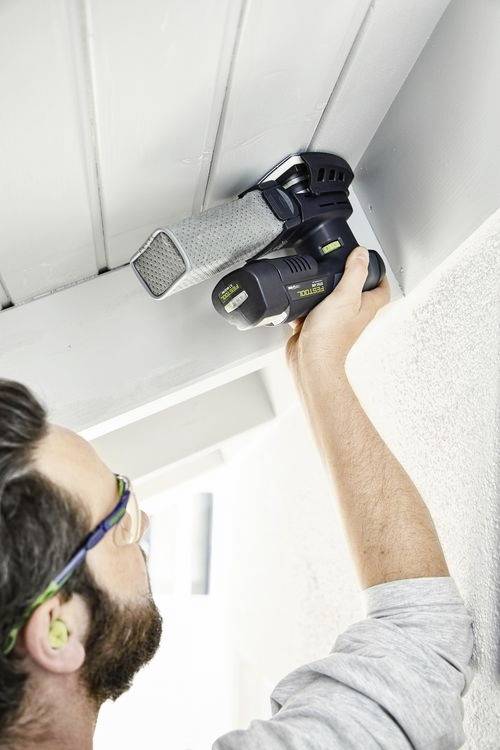 Eine Person, die mit einem tragbaren Elektrowerkzeug einen grauen Deckenbalken schleift, trägt Schutzbrille und Ohrstöpsel in einer hellen, Innenraum-Umgebung.