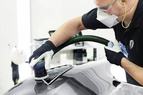 Eine Person mit Sicherheitsausrüstung poliert ein Automobilbauteil mit einer Poliermaschine und demonstriert dabei Fahrzeugaufbereitung in einer Werkstattumgebung.