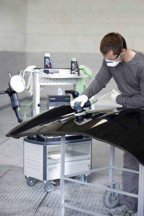 Eine Person mit Maske und Handschuhen benutzt eine Maschine, um die Motorhaube eines schwarzen Autos in einer Werkstattumgebung zu polieren.