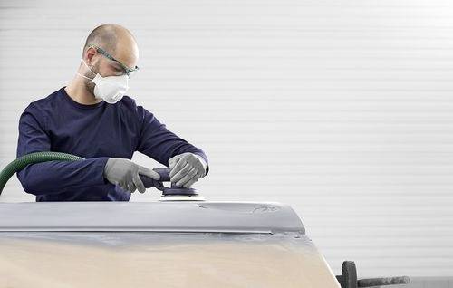Eine Person mit Schutzausrüstung benutzt ein Schleifwerkzeug auf der Oberfläche eines Fahrzeugs und konzentriert sich auf einen bestimmten Bereich in einer Werkstattumgebung.