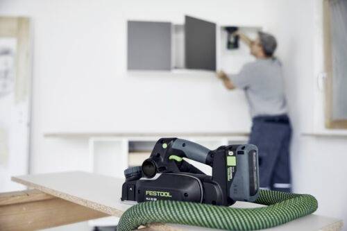 Eine kabellose elektrische Werkzeug mit einem Schlauch liegt auf einem Tisch; eine Person im Hintergrund montiert Regale an einer weißen Wand.