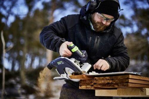 Eine Person, die eine Kreissäge verwendet, um Holzbretter im Freien zu schneiden, trägt Schutzausrüstung vor einer Hintergrund von Bäumen.