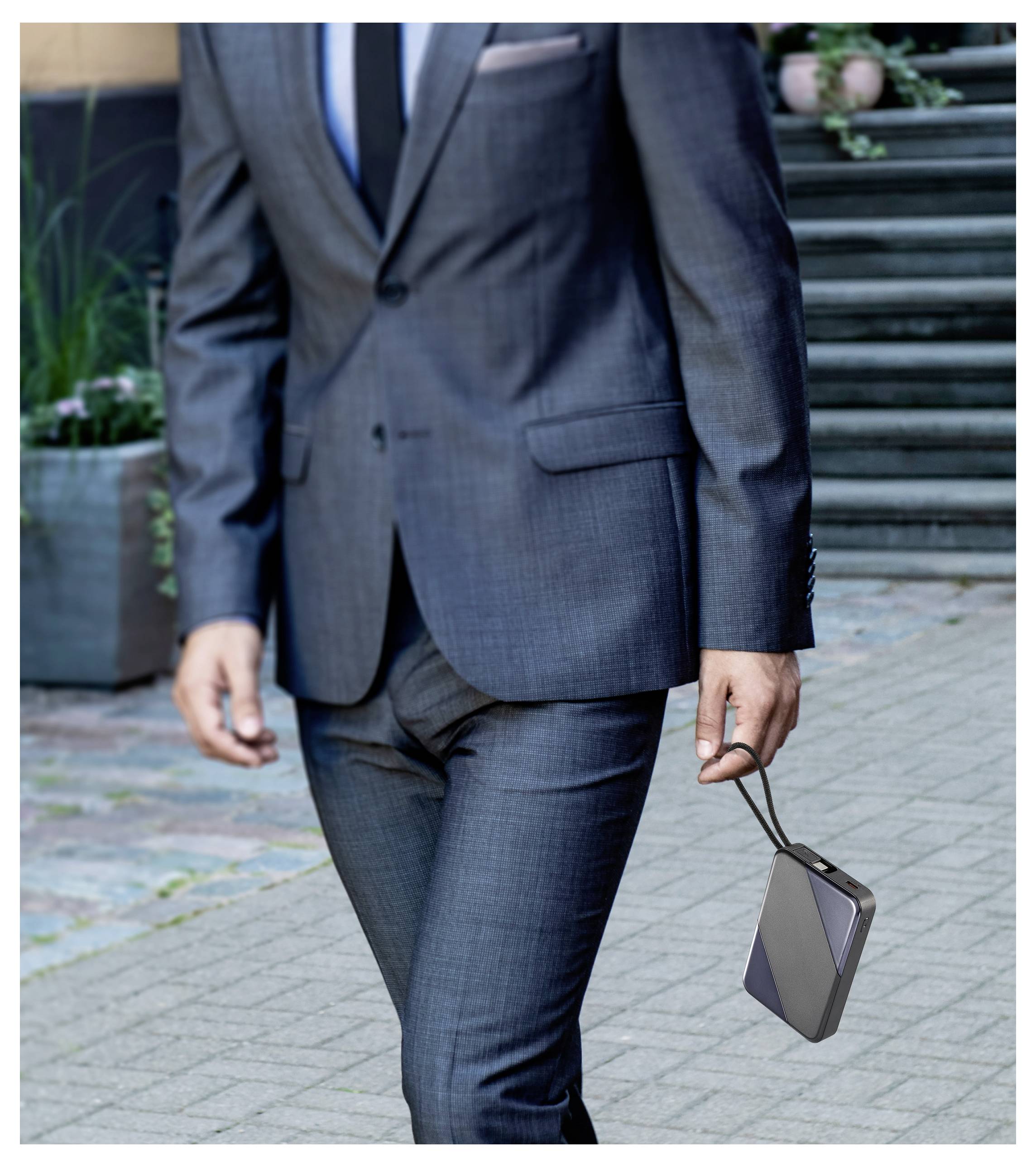 Eine gut gekleidete Person im Anzug geht nach draußen, während sie ein elegantes tragbares elektronisches Gerät an einem Riemen hält.