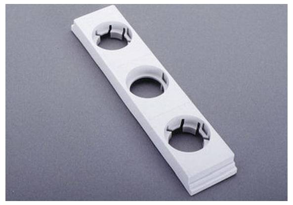 Ein weißes, rechteckiges Plastikpaneel mit drei kreisförmigen Öffnungen, die jeweils mit Clips versehen sind, liegt auf einer dunkelgrauen Oberfläche.