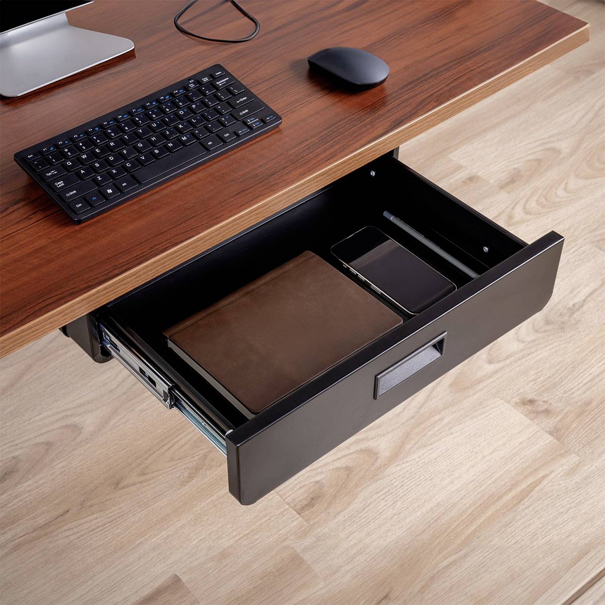 Eine offene Schublade gibt einen geschlossenen Laptop, ein braunes Notizbuch und ein Tablet im Inneren preis. Eine Tastatur und eine Maus befinden sich auf dem Schreibtisch darüber.