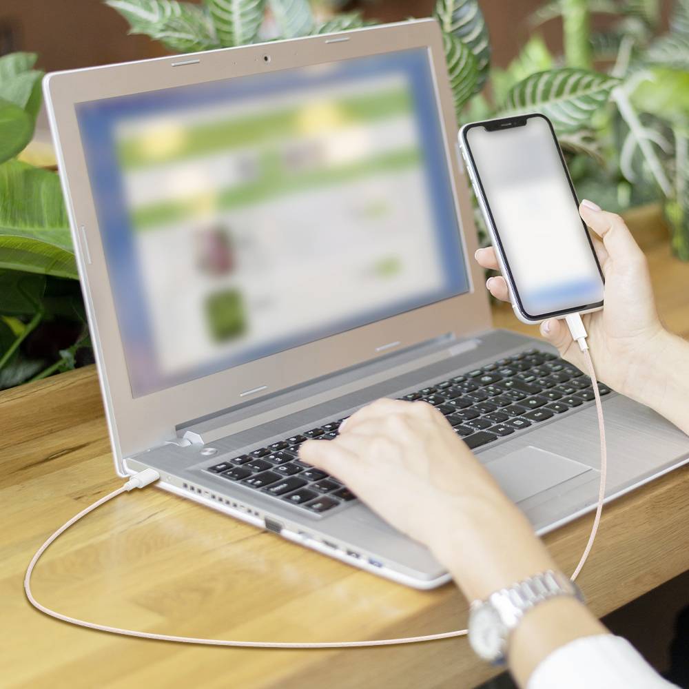 Eine Person, die mit einem Laptop interagiert und dabei ein Smartphone hält, wobei beide Geräte durch ein Kabel miteinander verbunden sind und damit eine Datenübertragung oder Synchronisation andeuten.