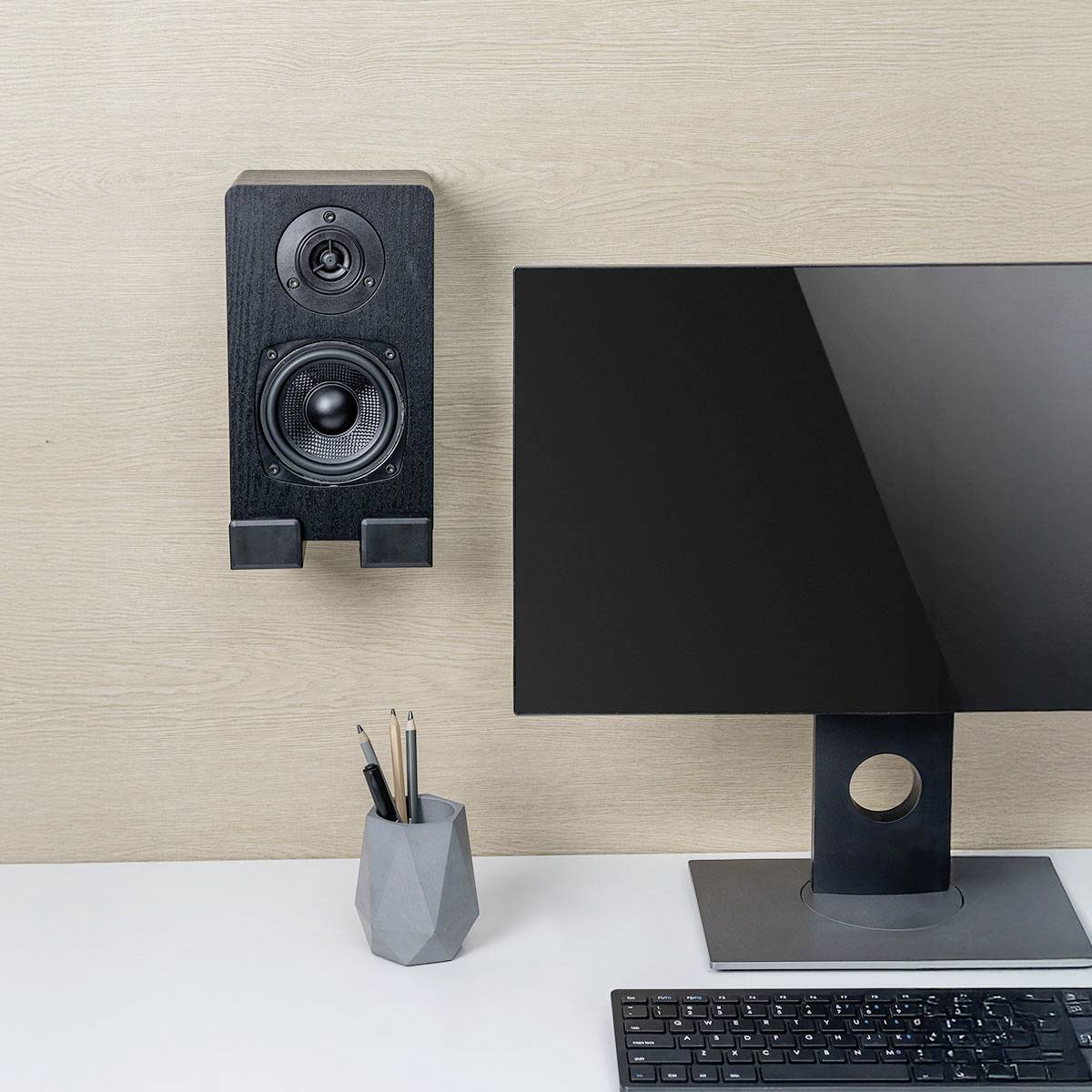 Ein Arbeitsplatz mit einem Computermonitor auf einem Ständer, einer an der Wand montierten Lautsprecherbox und einem Stiftehalter mit Stiften, der auf einem weißen Schreibtisch steht.