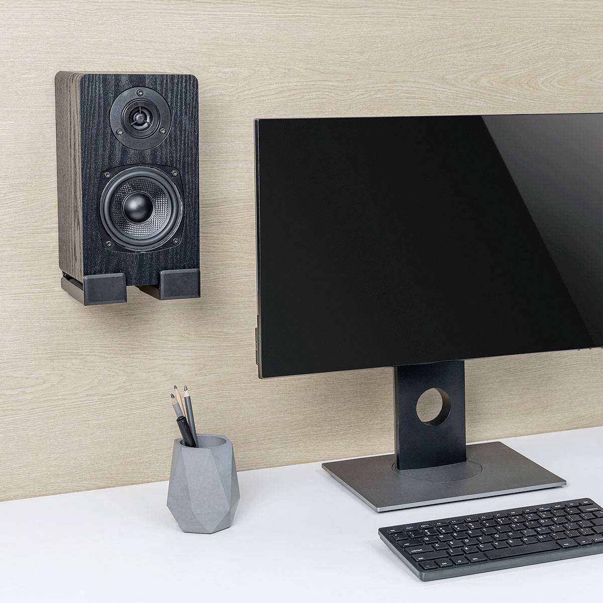 Ein Computermonitor auf einem Ständer, ein rechteckiger Stiftehalter mit Stiften und ein an der Wand befestigter Lautsprecher vor einem weiß-holzfarbenen Hintergrund.