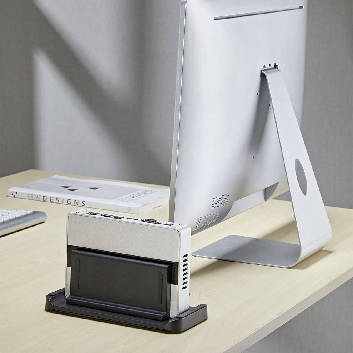 Ein kleiner Computer, der hinter ihm auf einem Schreibtisch an einen Monitor angeschlossen ist, mit einem Buch mit dem Titel „Great Designs", das in der Nähe positioniert ist.