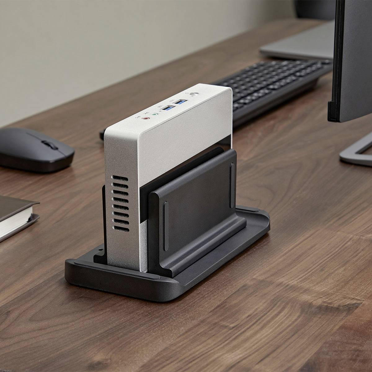 Ein Desktop verfügt über eine silberne Dockingstation mit verschiedenen Anschlüssen an der Seite, neben einer schwarzen Tastatur und Maus auf einem Holzschreibtisch.