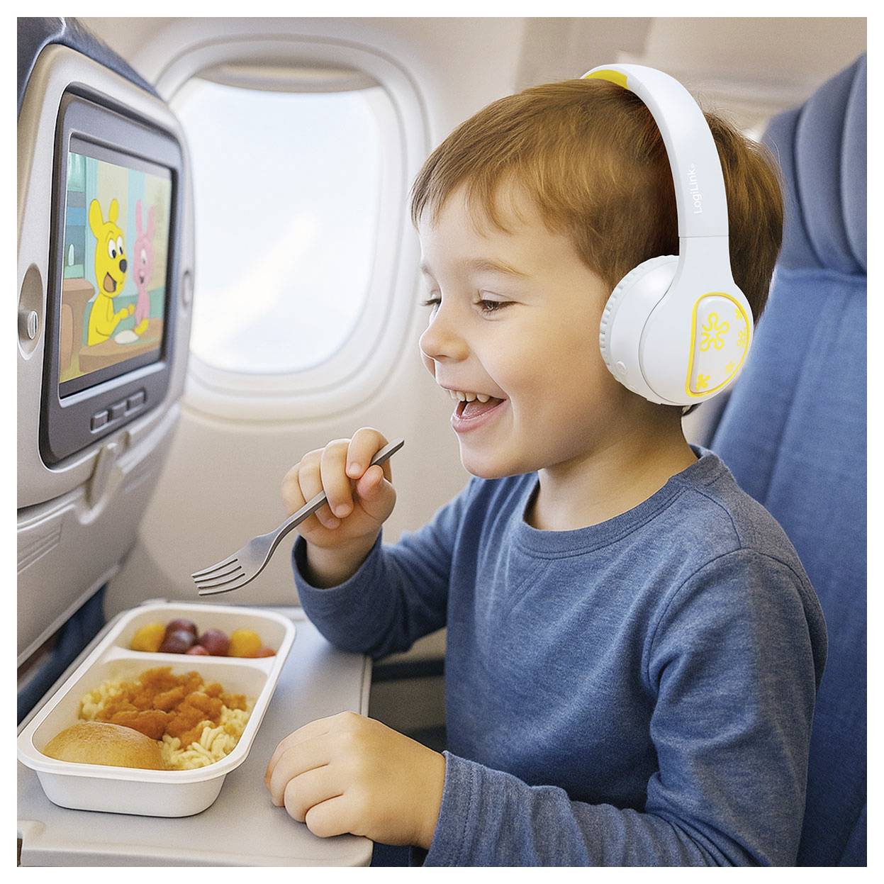 Ein Kind in einem Flugzeug, mit Kopfhörern, lächelt, während es einen Cartoon auf dem Sitzbildschirm anschaut. Es isst eine Mahlzeit von einem Essentablett.