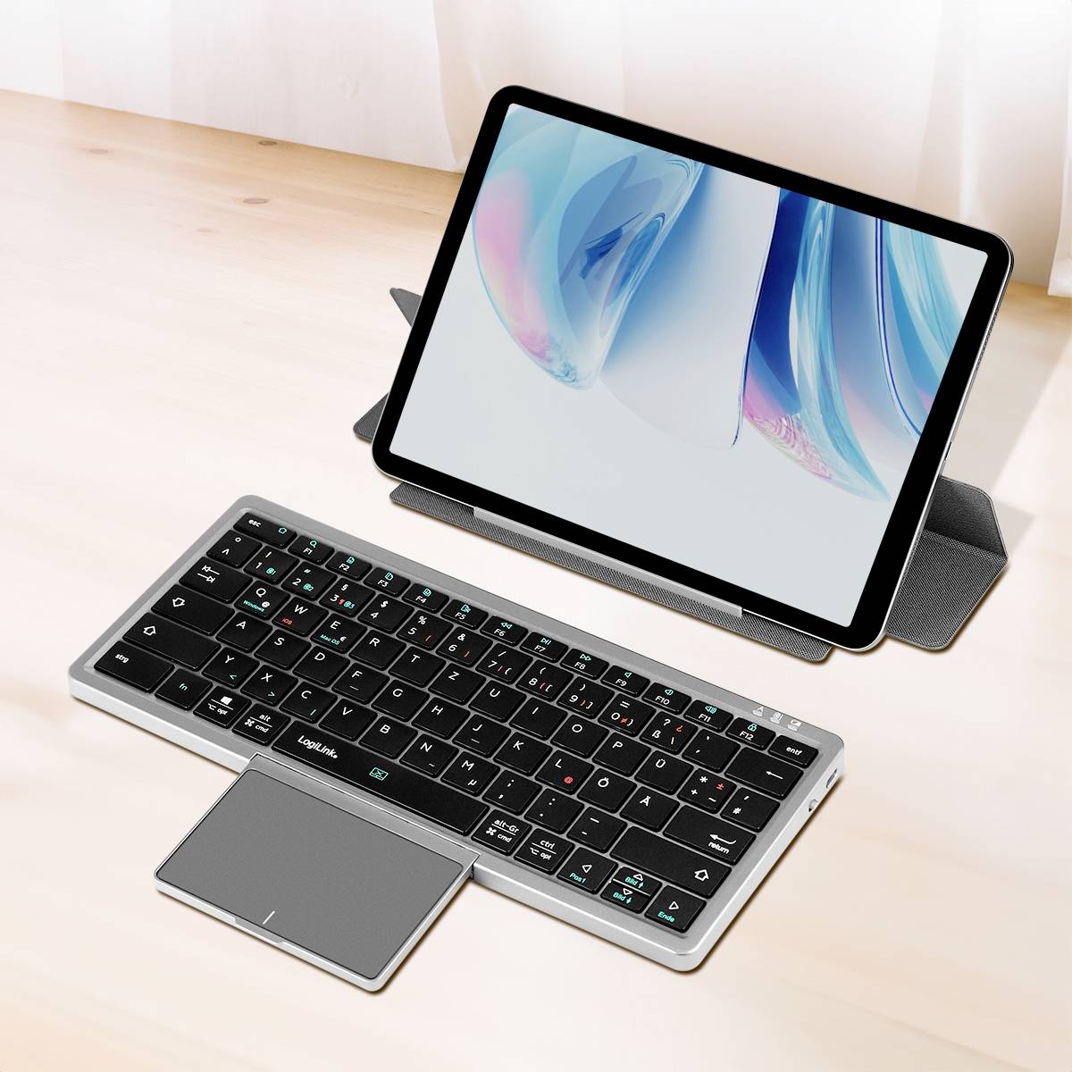 Ein Tablet steht auf einer Tasche hinter einer drahtlosen Tastatur mit Touchpad auf einem Holzschreibtisch und zeigt ein helles abstraktes Design auf dem Bildschirm.