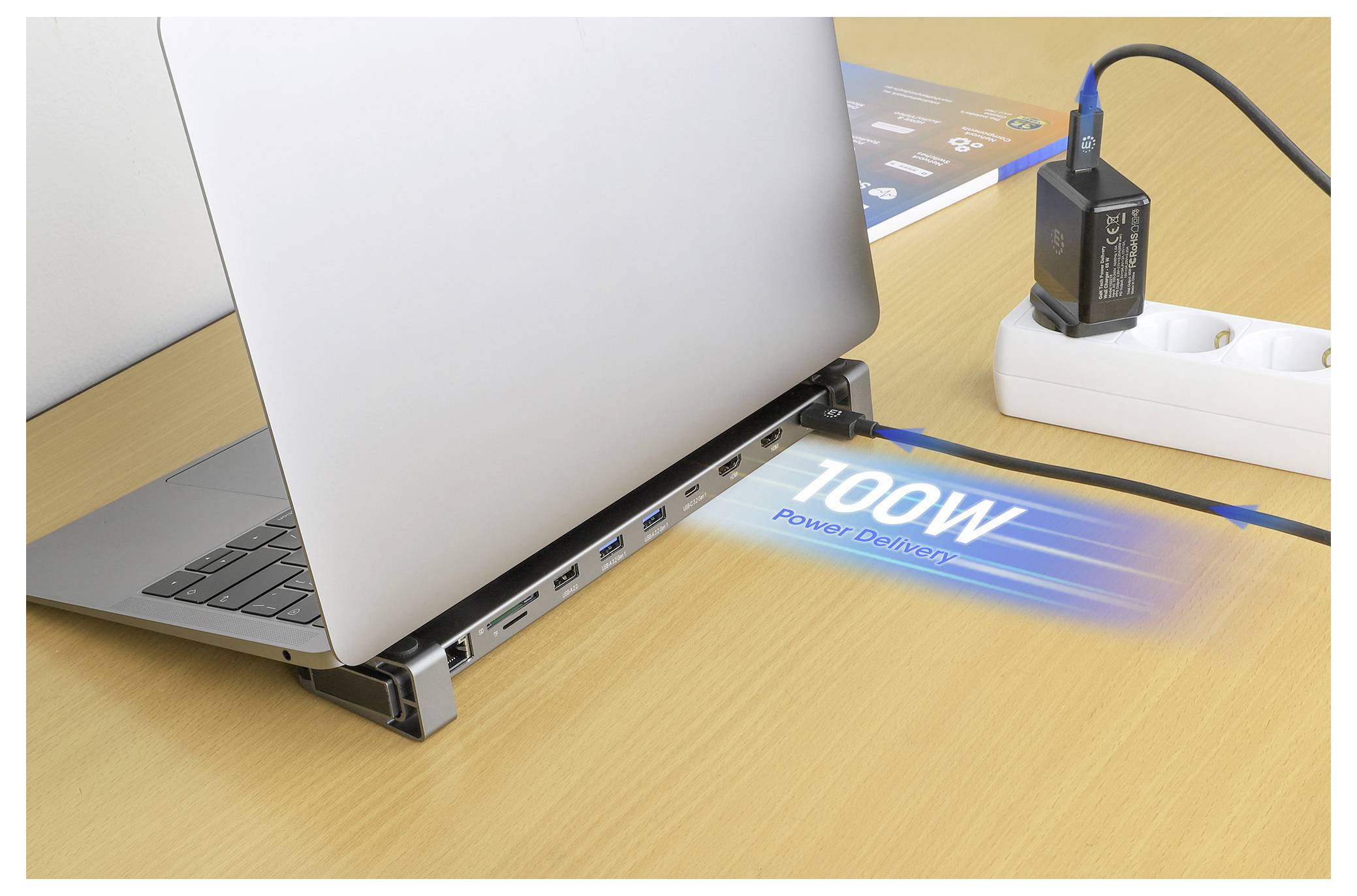 Ein Laptop ruht auf einer Dockingstation, auf dem Schreibtisch wird '100W Power Delivery' angezeigt und betont damit die hohe Ladekapazität. Ein Mehrfachstecker mit einem eingesteckten Adapter ist zu sehen.