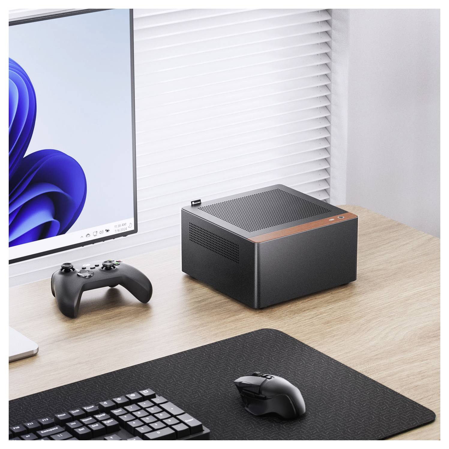 Ein kompakter schwarzer und orangefarbener Mini-PC steht auf einem Holzschreibtisch neben einem Computermonitor, der ein blaues abstraktes Logo und einen Gamecontroller zeigt.