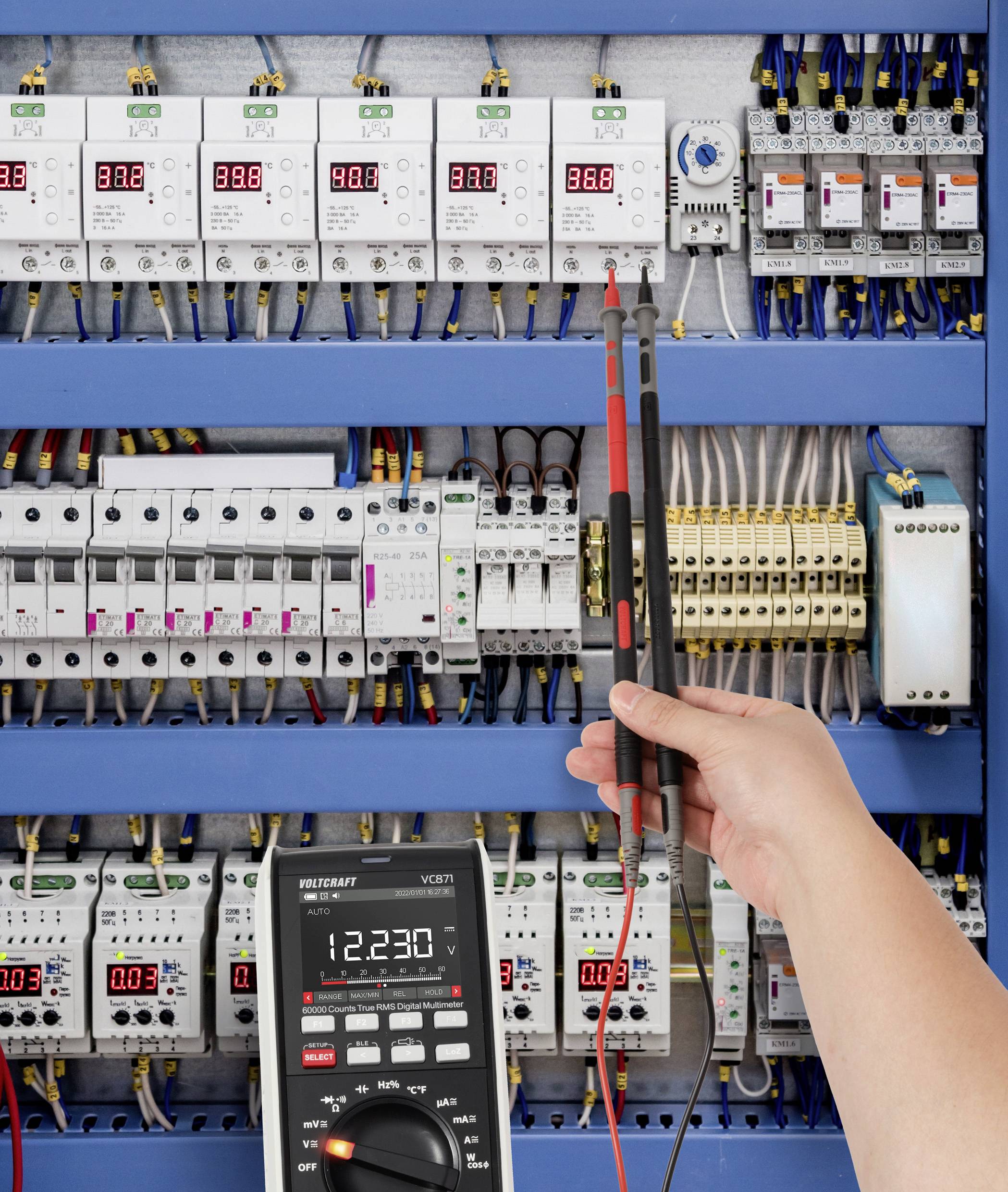 Eine Hand hält Multimeter-Messspitzen, die ein elektrisches Panel messen, mit verschiedenen digitalen Anzeigen, die Messwerte um die 380 Volt zeigen.