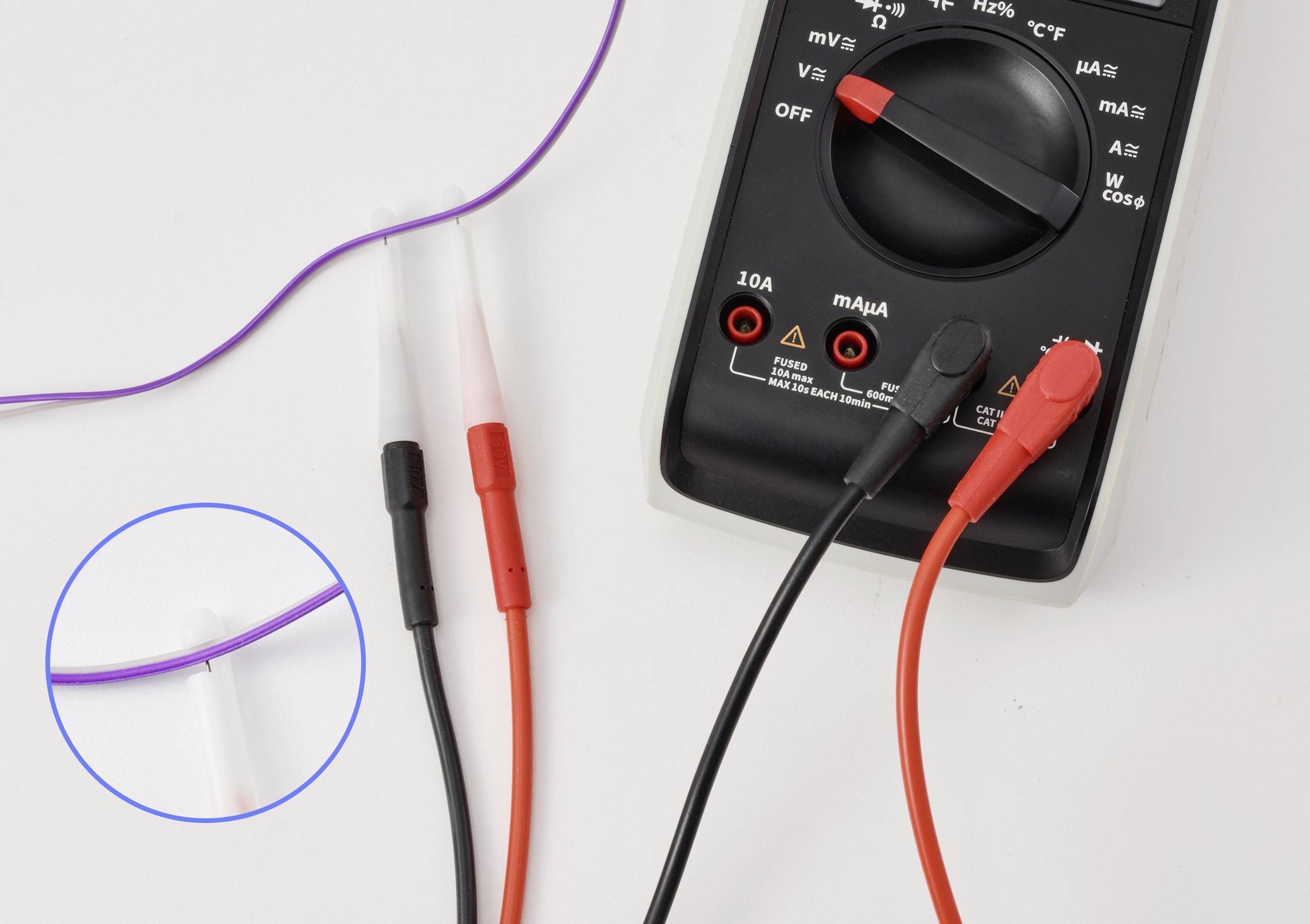 Ein digitales Multimeter ist zu sehen, mit roten und schwarzen Messleitungen, und ein Ausschnittbild hebt die Messspitzen hervor, die eine lila Leitung berühren.