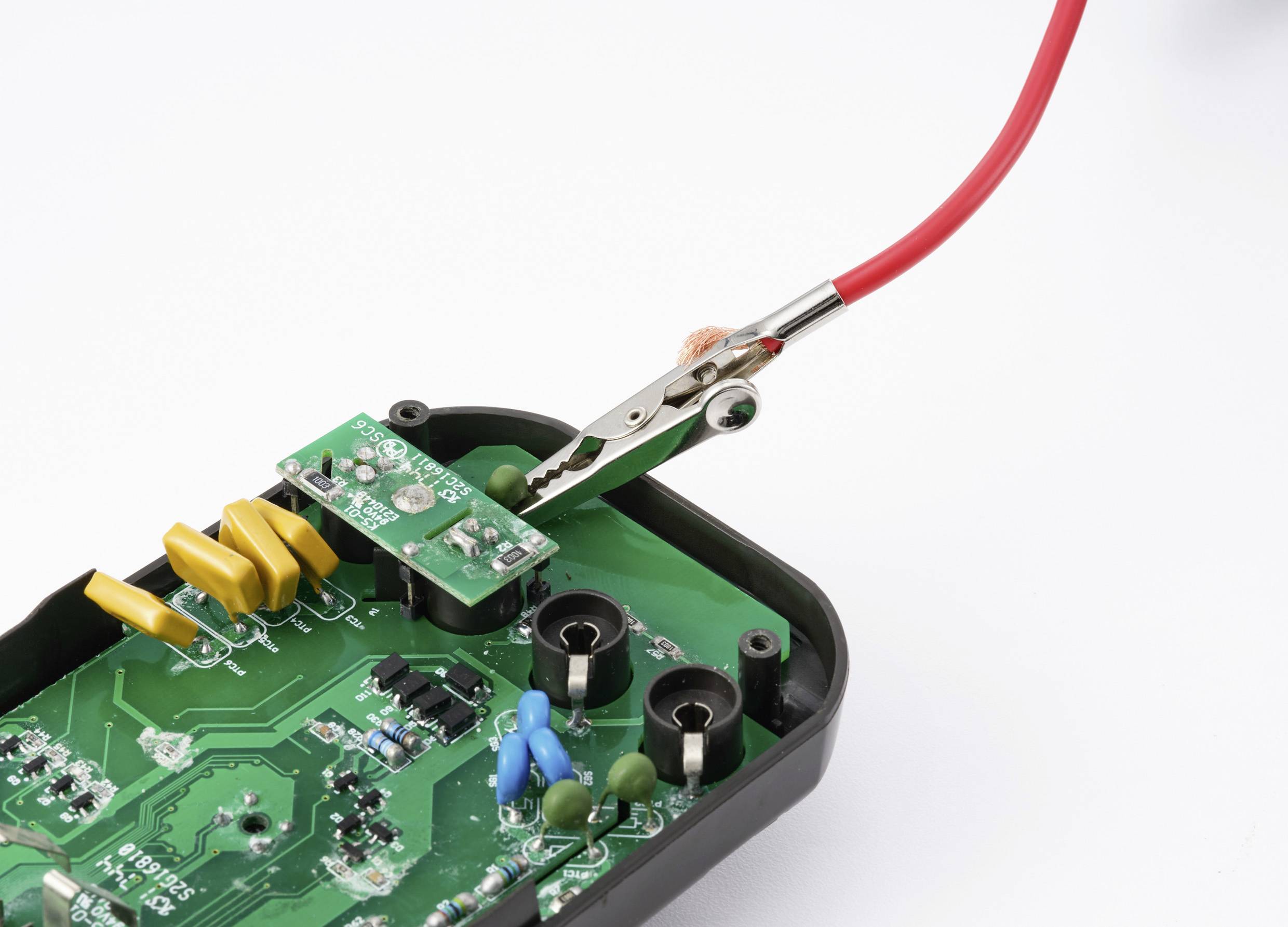 Eine Leiterplatte mit einem Testklemmer und einem roten Kabel, die kleine Komponenten und grüne Plattendetails zeigt, die für elektronische Tests verwendet wird.