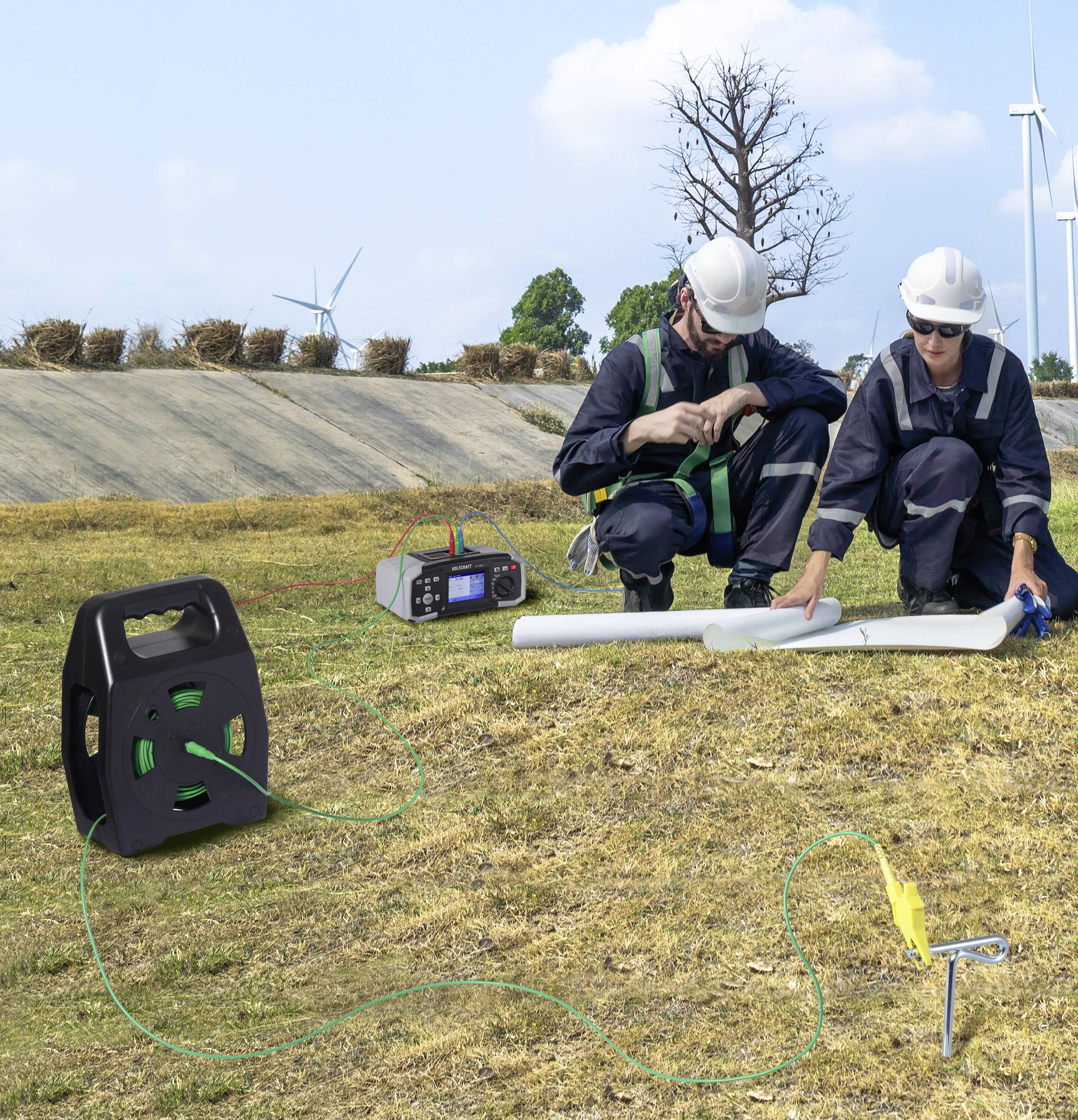 Zwei Arbeiter in Helmen und Sicherheitsausrüstung besprechen Pläne auf einem grasigen Feld mit Windturbinen. Ausrüstung liegt in der Nähe, einschließlich eines Generators.