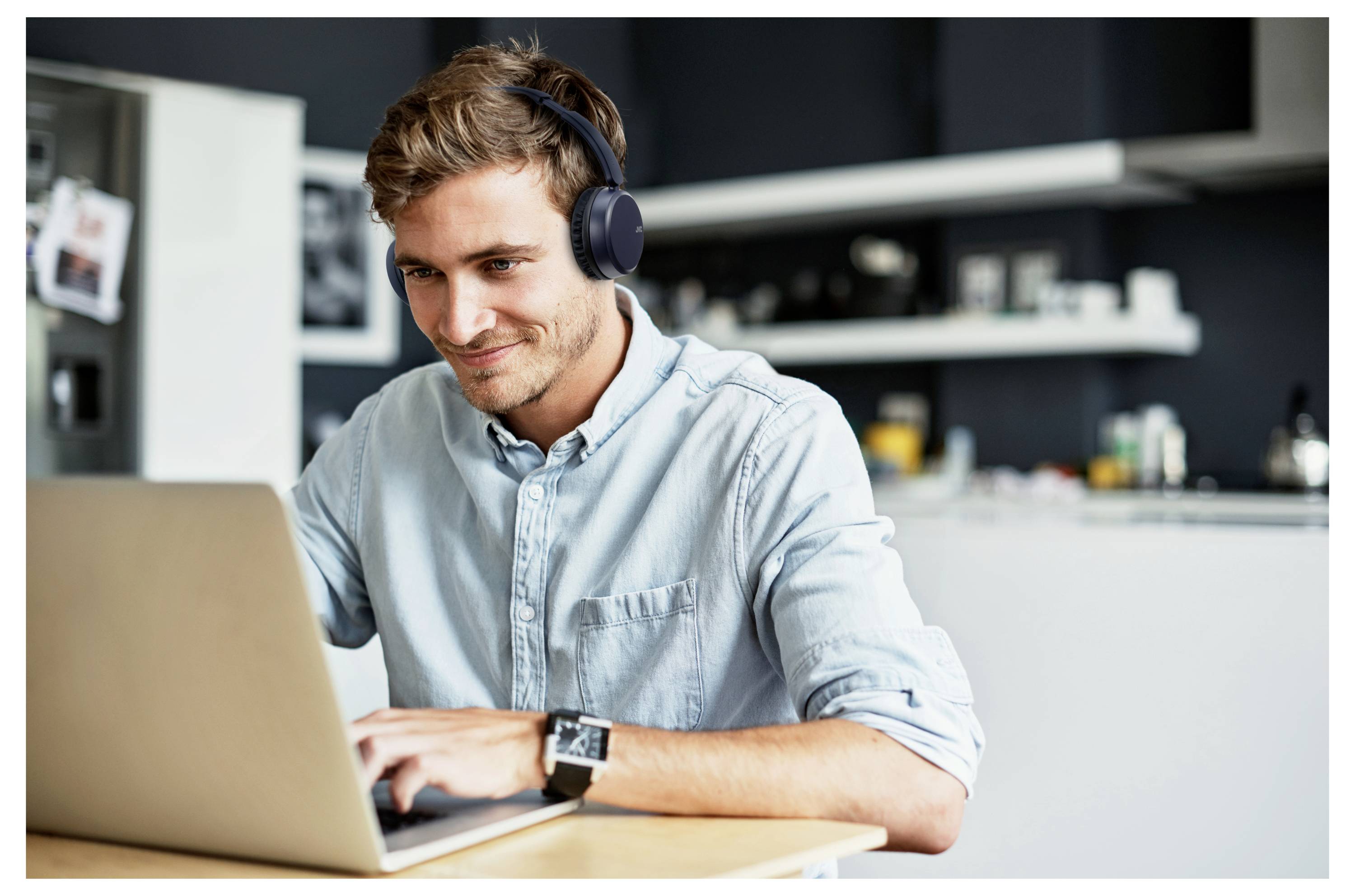 Ein Mann in einem lässigen Hemd sitzt an einem Tisch, arbeitet an einem Laptop und trägt Kopfhörer in einer modernen Küchenumgebung.