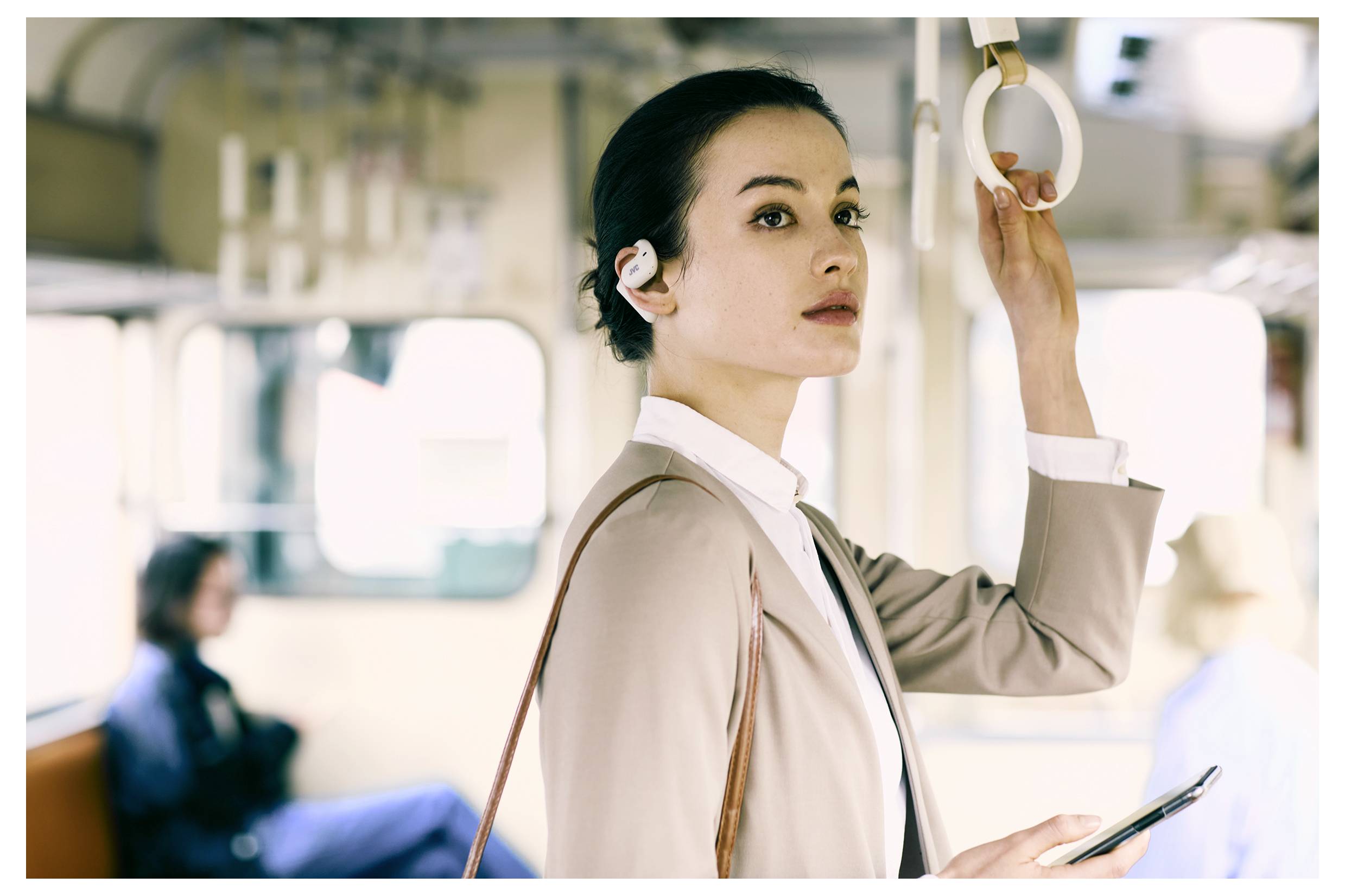 Eine Person in Geschäftskleidung hält sich in einem fahrenden Zug an einem Halteriemen fest und blickt nach vorn. Sie trägt Ohrhörer und hält ein Smartphone in der Hand.