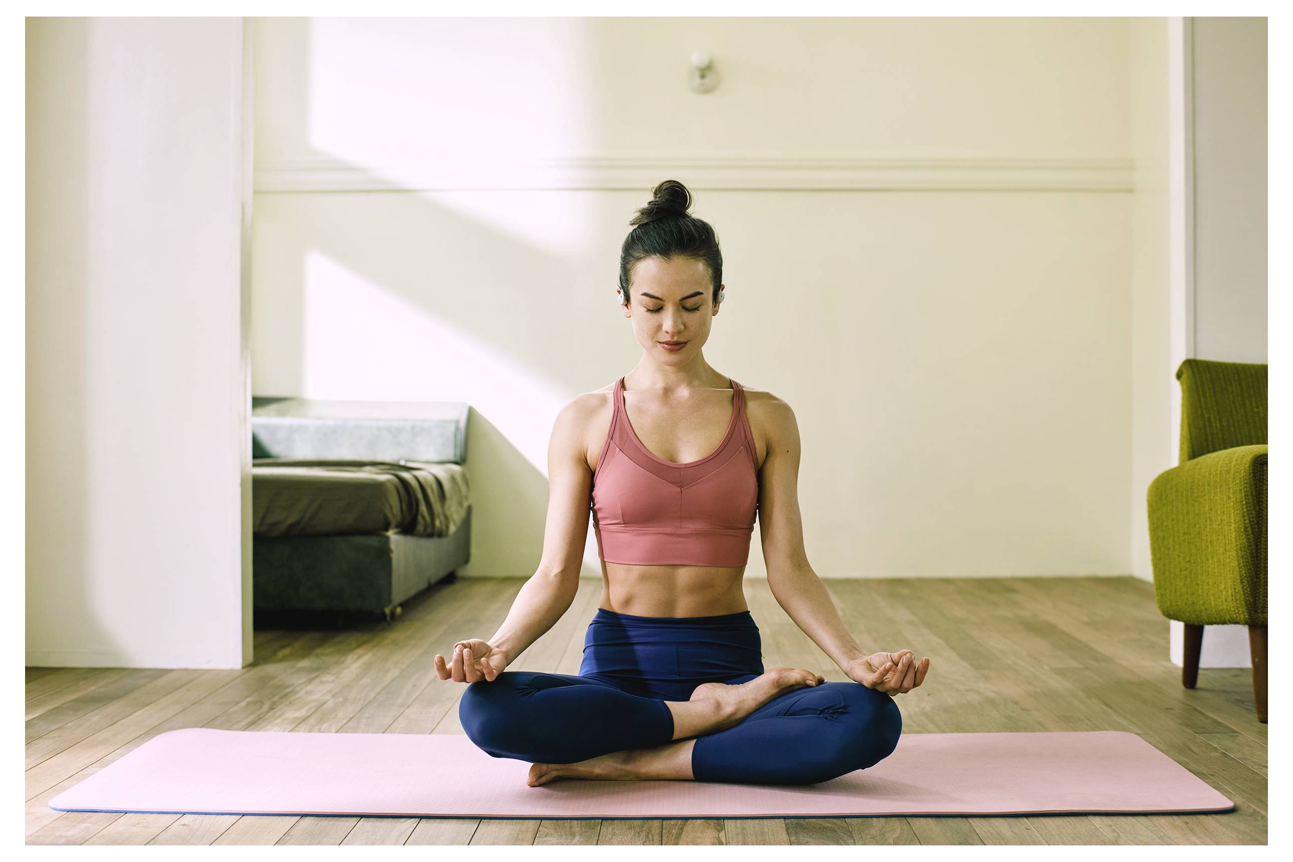 Eine Person praktiziert Yoga in einem ruhigen Raum und sitzt mit verschlossenen Augen im Schneidersitz auf einer rosa Matte, trägt ein rosa Oberteil und blaue Hose.