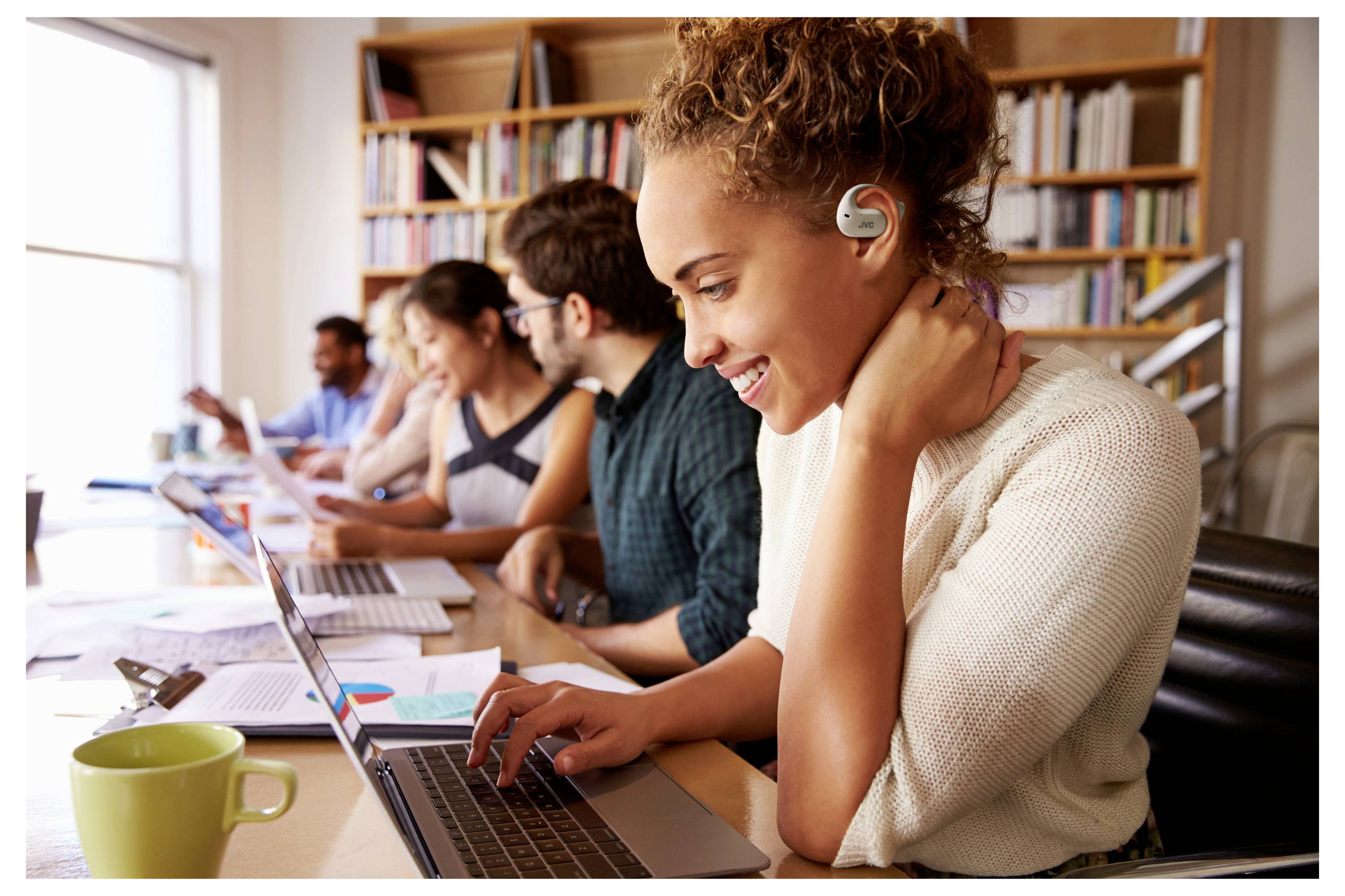 Eine Frau mit einem Hörgerät lächelt, während sie in einer Bibliothek einen Laptop benutzt. Im Hintergrund arbeiten mehrere andere Personen an Laptops.