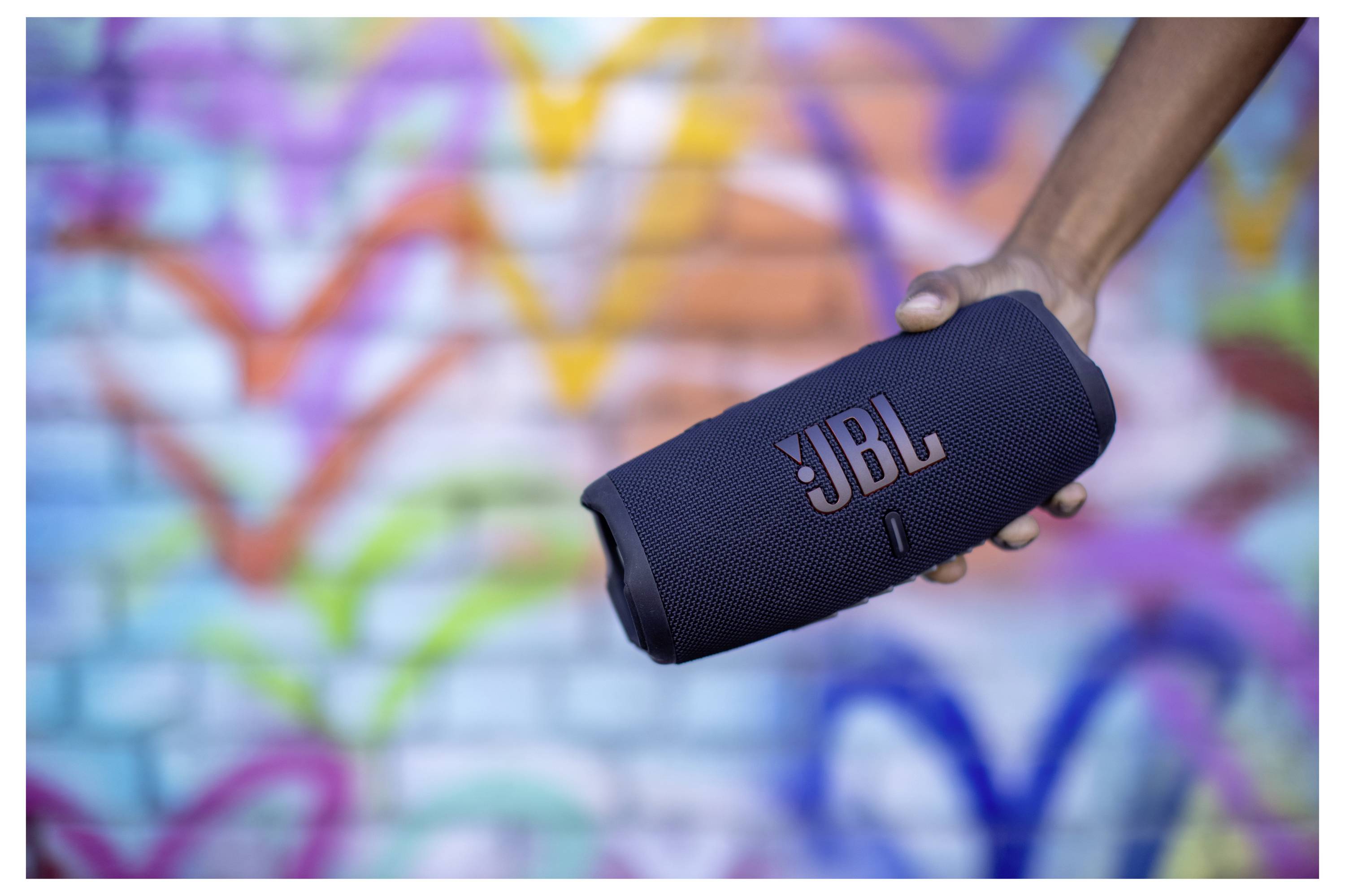 Eine Person hält einen tragbaren JBL-Lautsprecher vor einer bunten Graffiti-Wand und demonstriert damit die Mobilität des Lautsprechers vor einem lebhaften Designhintergrund.