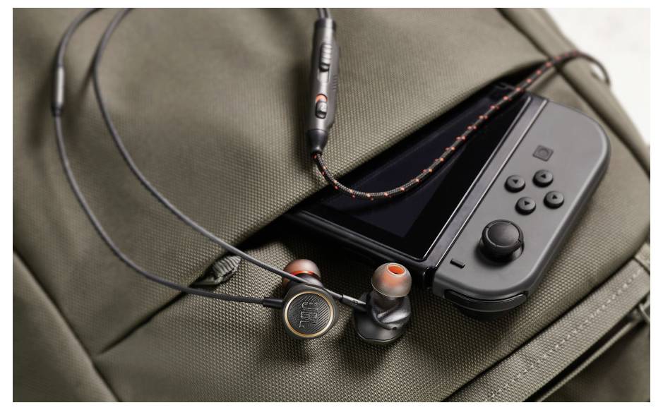 Eine Handheld-Spielkonsole und Kopfhörer, die teilweise in der vorderen Tasche eines geschlossenen, grauen Rucksacks stecken und Reisen oder Mobilität andeuten.