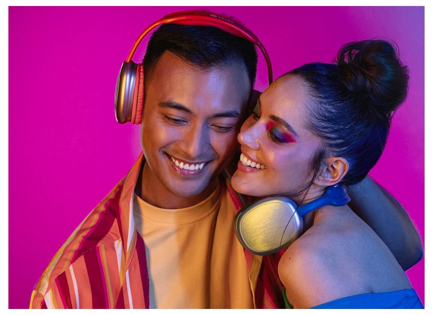 Ein Mann und eine Frau genießen gemeinsam Musik, tragen Kopfhörer und lächeln vor einem lebendigen rosa-lila Hintergrund.