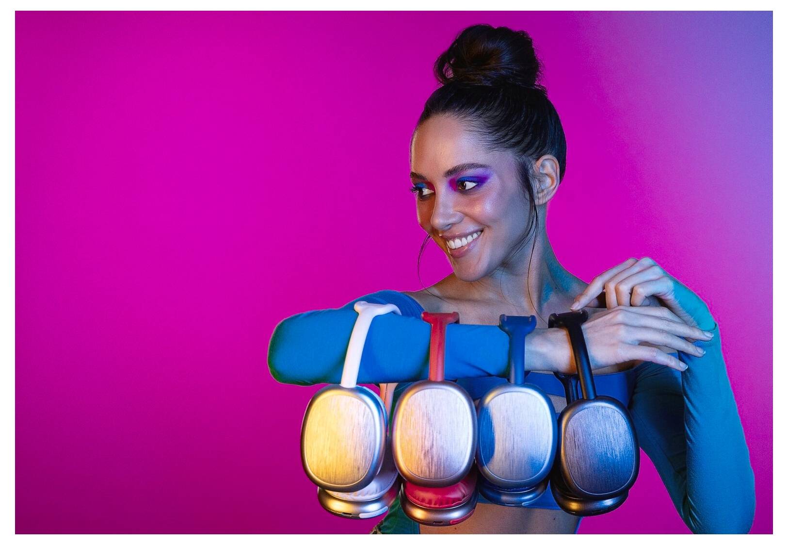 Eine Frau in einem bunten Outfit posiert vor einem pinkfarbenen Hintergrund, lächelt und hält fünf Kopfhörer-Paare in verschiedenen Farben.