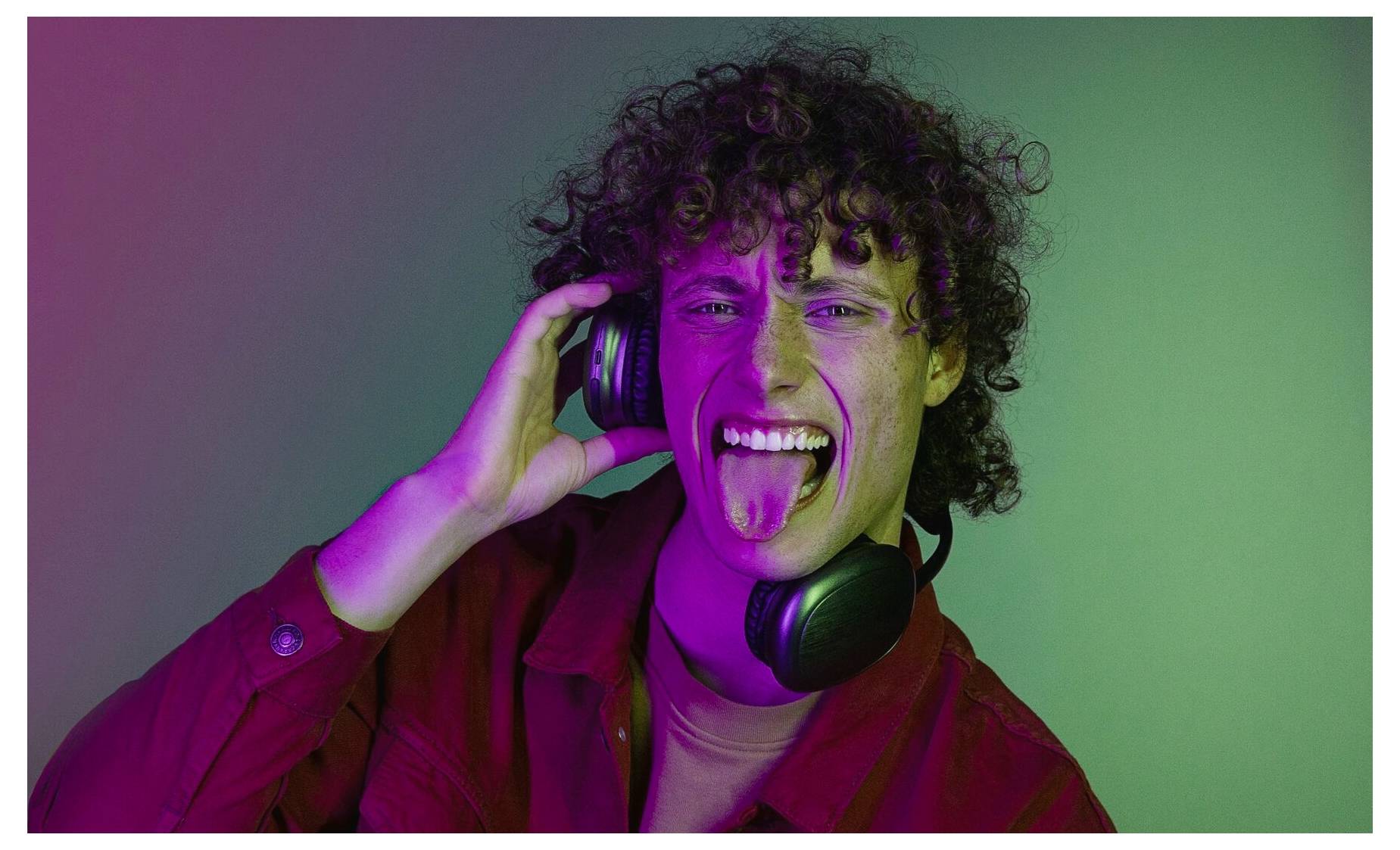 Eine Person mit Kopfhörern macht einen verspielten Gesichtsausdruck mit herausgestreckter Zunge vor einem bunten Hintergrund.