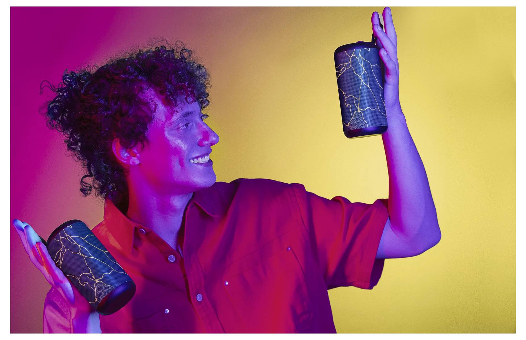 Eine Person lächelt und hält zwei gemusterte zylindrische Lautsprecher, während sie vor einem lebendigen Farbverlaufshintergrund mit gelben und rosa Tönen steht.