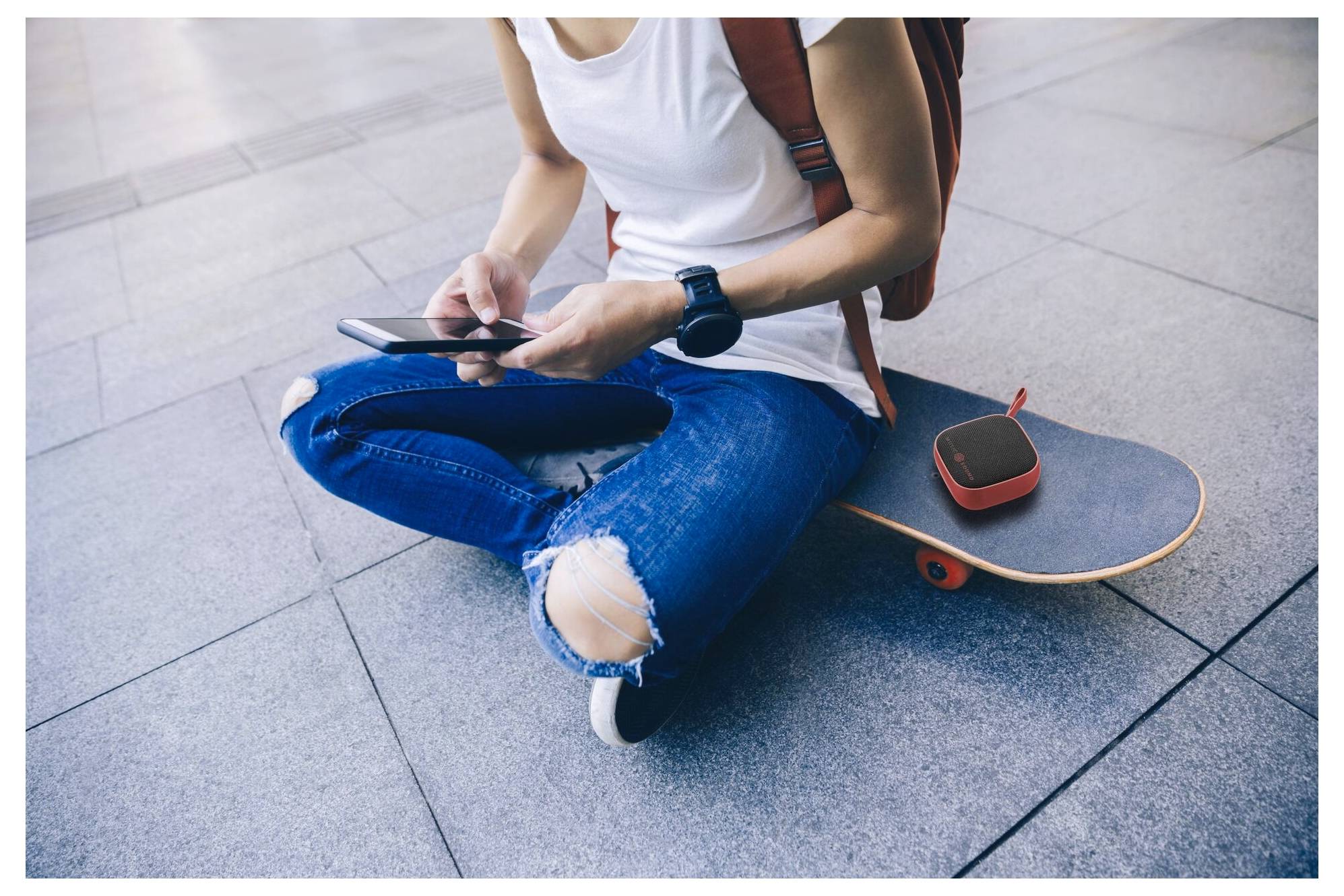 Eine Person, die mit gekreuzten Beinen auf dem Gehweg sitzt, mit einem Skateboard und einem tragbaren Lautsprecher in der Nähe, während sie ein Smartphone benutzt.