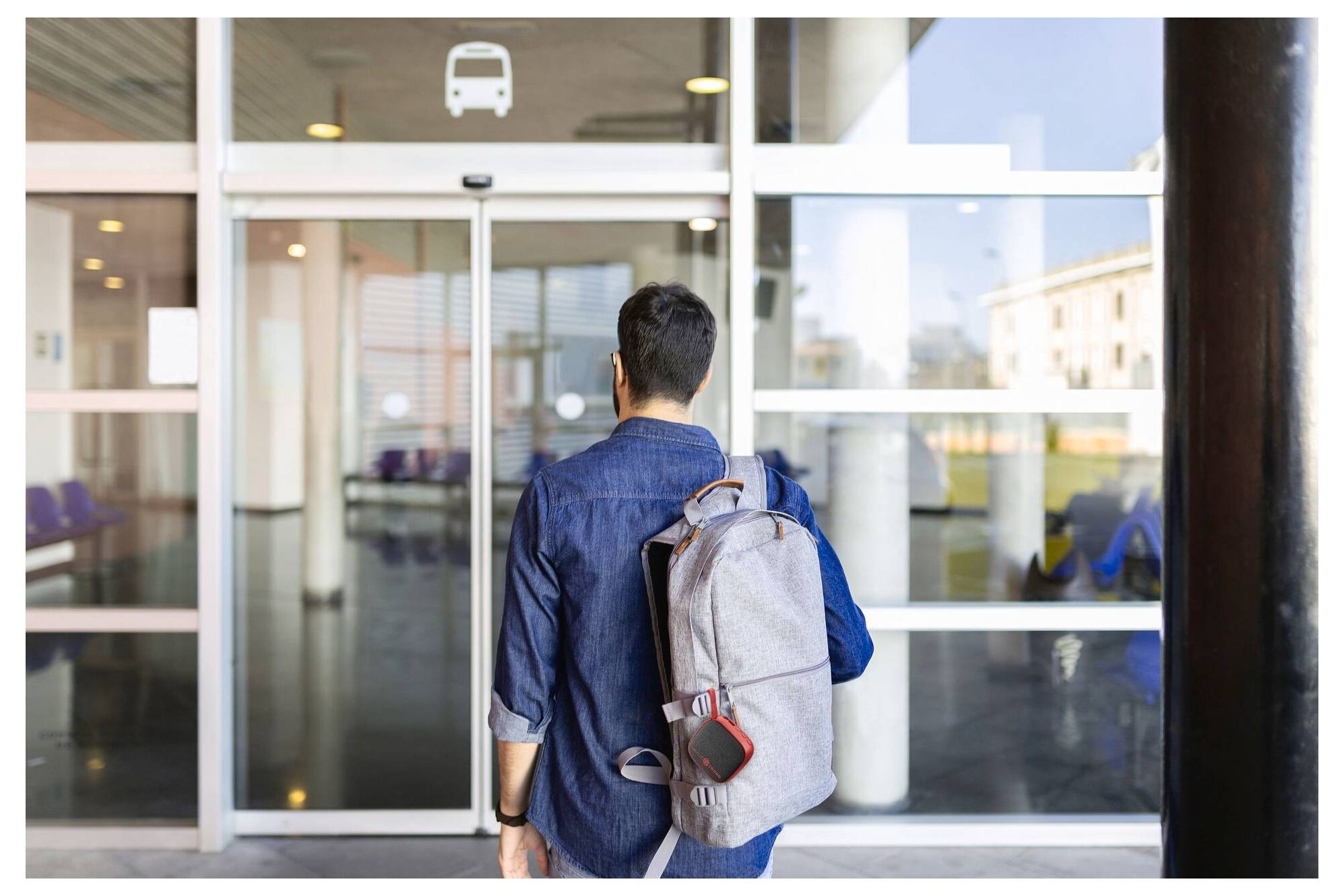 Eine Person mit einem Rucksack betritt eine Busstation durch eine Glastür, wobei Busikonen oben angezeigt werden.